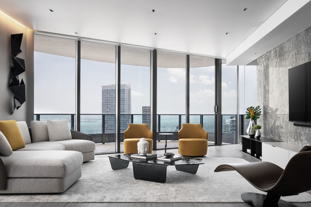 Modern living room with floor-to-ceiling windows, white boucle sectional sofa, mustard yellow armchairs, hammered black glass coffee table, sand rug, and city view.